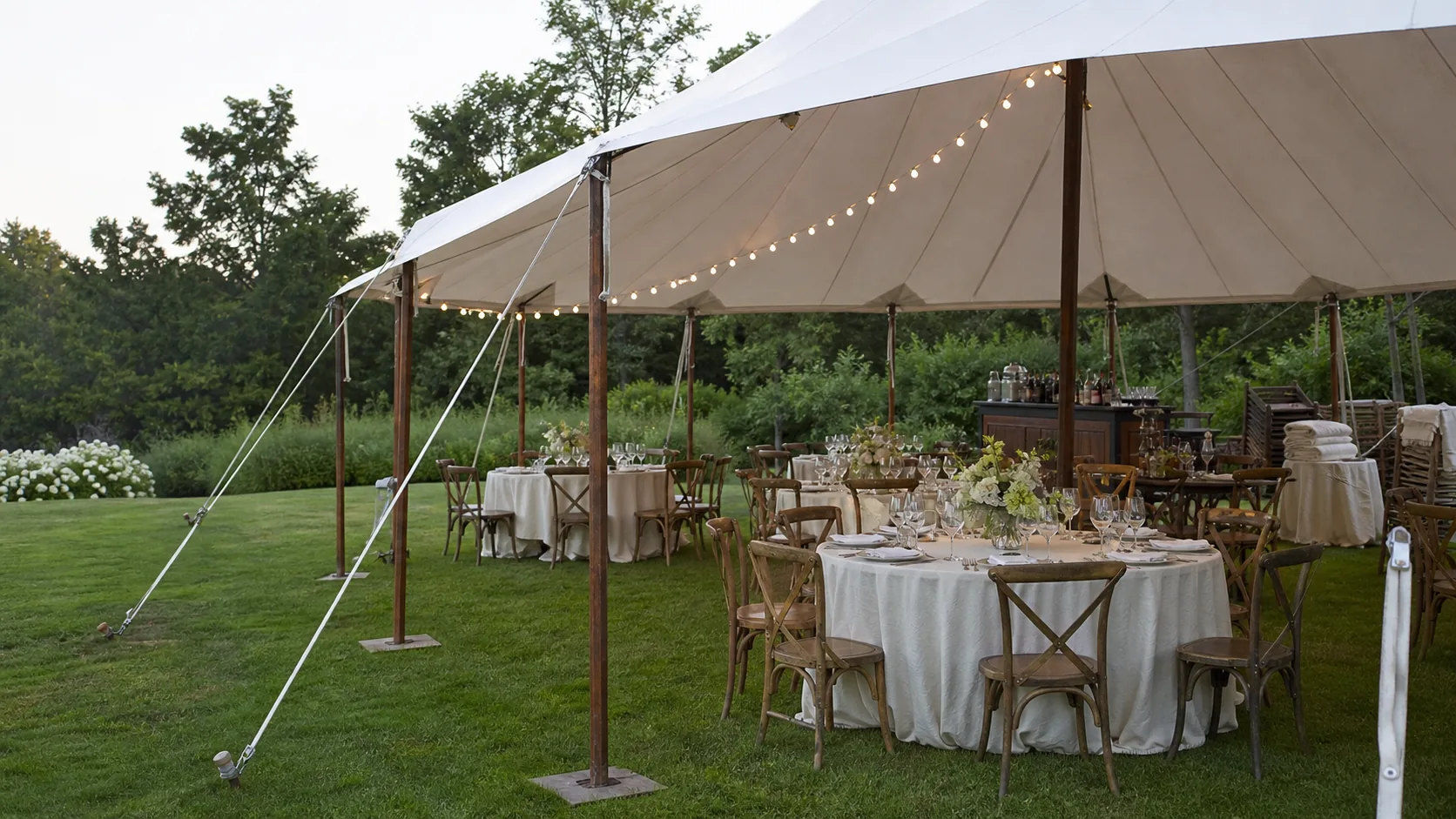 An outdoor event with a sailcloth tent, rental tables, wooden chairs, and linen table settings ready for guests.
